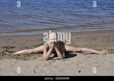 Bald donna seduta in split appoggiata al suo i gomiti su una sabbiosa spiaggia fluviale, sorridente Foto Stock