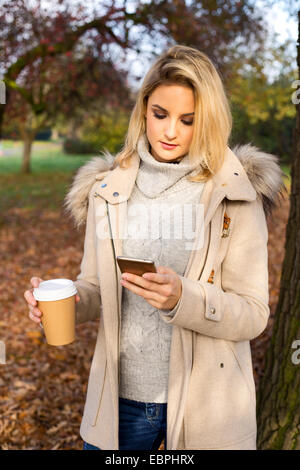 Bella giovane donna controlla i suoi messaggi con un caffè nel parco Foto Stock