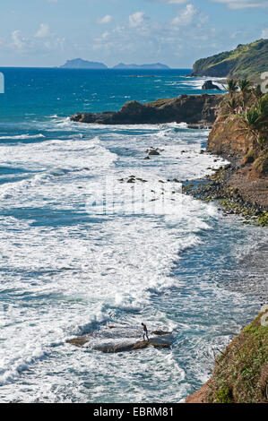 Vista lungo la costa di roccia con un pescatore in piedi su una roccia nel surf, Saint Vincent e Grenadine, Biabou Bay Foto Stock