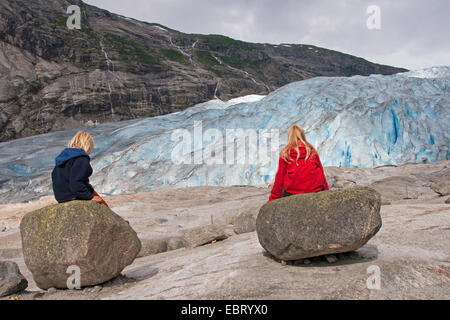 Due bambini seduti sulle rocce di fronte del ghiacciaio Nigardsbreen, Norvegia, Jostedalsbreen Parco Nazionale Foto Stock