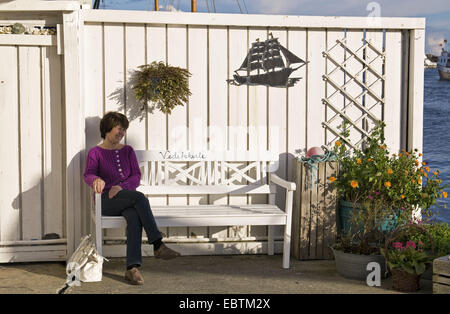 Donna seduta su una panchina del bianco nella parte anteriore del bianco recinzione in legno, Norvegia, Rogaland, Karmoy, Skudeneshavn Foto Stock