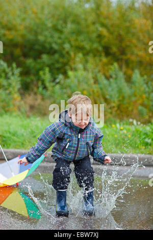 Little Boy jumping con stivali da pioggia in una pozza Foto Stock