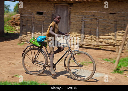 Little Boy in sella ad una bicicletta per adulti su una strada polverosa lungo fango umili case di mattoni, Burundi, Cankuzo, Nazionale Parc de la Ruvubu, Cankuzo Foto Stock