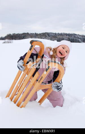 Bambina frolicly kneeing in un campo di neve con una pista da slittino in legno, Svizzera Foto Stock