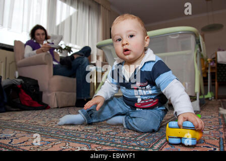 Little Boy giocando sul pavimento, sua madre seduta su un divano mantenendo un occhio su di lui Foto Stock