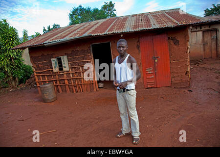 Giovane uomo è in piedi sulla parte anteriore della sua semplice casa di fango con un ferro corrugato tetto, Uganda, Jinja Foto Stock