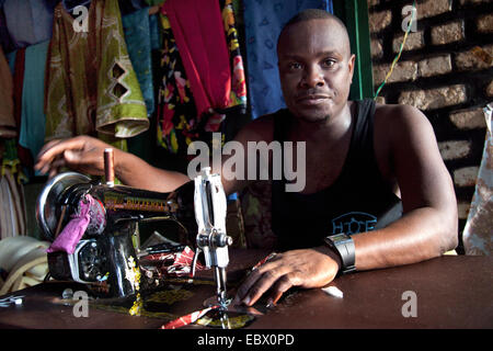 Tailer seduto alla macchina da cucire nel suo atelier, abiti finiti in background, Ruanda, Nyamirambo, Kigali Foto Stock