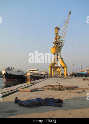 Lavoratore dorme nell'ombra sul terreno asfalto della banchina del porto di Bujumbura, grande gru e navi in background, Burundi Bujumbura marie, Bujumbura Foto Stock