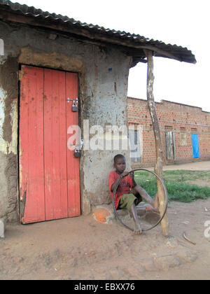 Ragazzo seduto accanto alla porta chiusa di un piccolo negozio con un vecchio pneumatico di una bicicletta in mani, Uganda, Gulu, John Baptist Odama Arcivescovo di Gulu Foto Stock