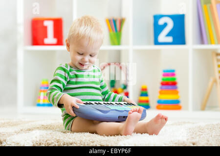 Felice capretto piccolo bimbo boy divertendosi pianoforte giocattolo seduto sul pavimento in vivaio Foto Stock