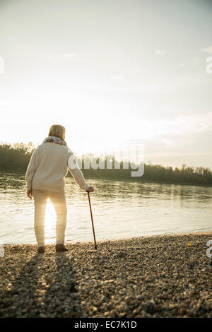 Senior donna con bastone da passeggio permanente al waterside guardando il tramonto, vista posteriore Foto Stock