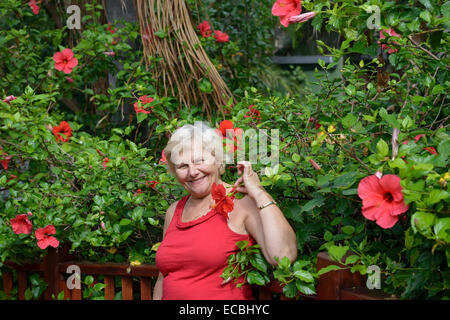 Vita ritratto di donna di età e su sfondo tropicale con fiori di colore rosso di Hibiscus bush. Foto Stock