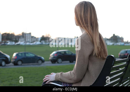 Vista posteriore del colpo di una giovane donna seduta su una panchina, esaminando la distanza in un parco con le automobili parcheggiate. Foto Stock