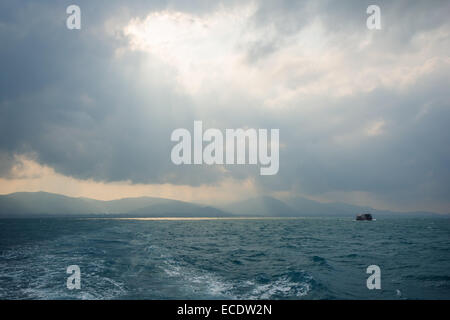 Raggi di sole attraverso il foro tra le nuvole sopra l'oceano e una nave, silhouette collinari del Koh Samui, Thailandia Foto Stock