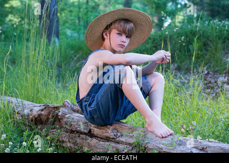 Ragazzo seduto sul log indossando cappello di paglia e tute blu Foto Stock