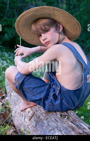 Ragazzo giovane indossando tute seduta su albero indossando cappello di paglia Foto Stock