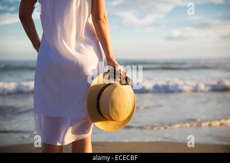 Vista posteriore della donna in abito bianco permanente sulla spiaggia e azienda hat Foto Stock