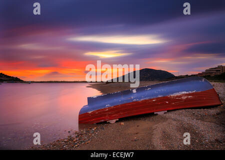 Lonely barca da pesca a sinistra a riva contro uno spettacolare e colorato tramonto con nubi colorate che passa al di sopra della barca Foto Stock