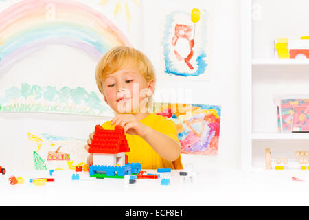 Poco tedesco cerca ragazzo biondo con riproduzione di materia plastica in blocchi di costruzione semplice casa nella stanza dei bambini setup Foto Stock