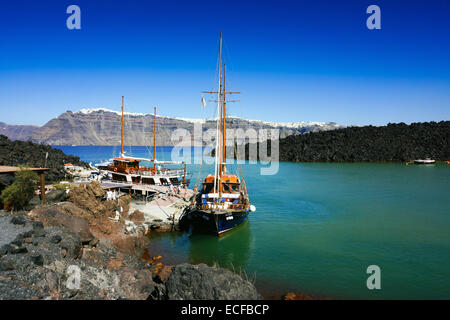 Pontile con imbarcazioni turistiche Nea Kameni lava vulcanica isola, Santorini, Grecia, con Fira sul bordo del cratere. Foto Stock