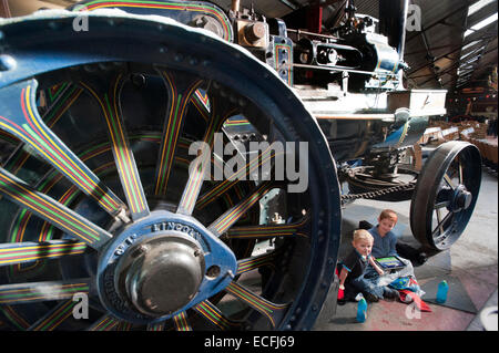 Due giovani fratelli sedersi sotto le gigantesche ruote decorativo del motore di trazione "il campione" costruito da Ruston Proctor & Co di Lincoln in sala Strumpshaw Steam Museum vicino a Norwich, Norfolk. Il motore di trazione è stato sul display insieme con rulli a vapore, motori showmans e motori di aratura in exhibition hall. Foto Stock