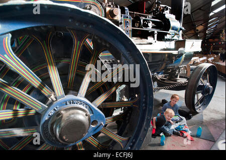 Due giovani fratelli sedersi sotto le gigantesche ruote decorativo del motore di trazione "il campione" costruito da Ruston Proctor & Co di Lincoln in sala Strumpshaw Steam Museum vicino a Norwich, Norfolk. Il motore di trazione è stato sul display insieme con rulli a vapore, motori showmans e motori di aratura in exhibition hall. Foto Stock