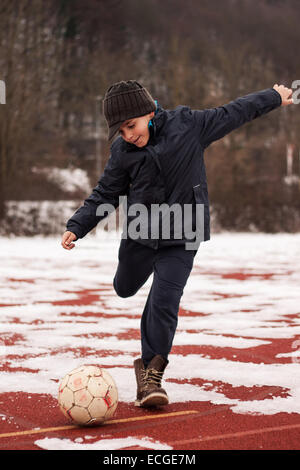 Ragazzo calciare il pallone nella stagione invernale sul posto rosso con lastre di ghiaccio Foto Stock