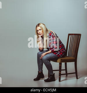 Studio shot della giovane donna in abito casual seduti su una sedia guardando la fotocamera. Caucasian modello femminile con copyspace. Foto Stock