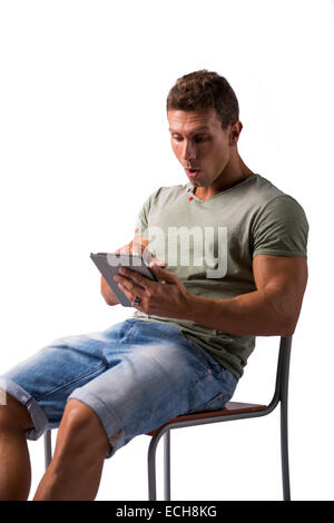 Muscolare di giovane uomo seduto su una sedia mentre la lettura da e-book dispositivo con espressioni di sorpresa, isolato su bianco Foto Stock