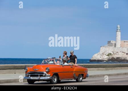 Cuba, La Habana, turisti che si siedono in una vettura americana sul Malecon e Castillo de los Tres Reyes Magos del Moro in background Foto Stock