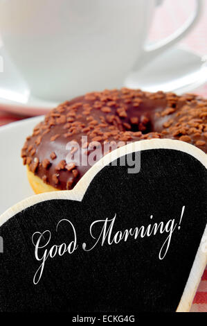 Primo piano di una a forma di cuore lavagna con la frase buona mattina scritto in esso e un cioccolato ciambella rivestito e una tazza di coff Foto Stock