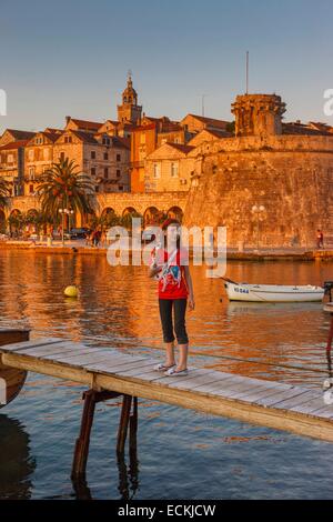 Croazia, Dalmazia, costa dalmata, Isola di Korcula Korcula, una giovane ragazza in piedi su un ponte di legno di fronte ad una porta fortificata città al tramonto Foto Stock