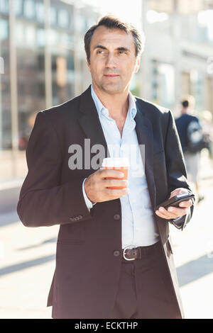 Imprenditore che guarda lontano mentre si tiene il telefono cellulare usa e getta e tazza di caffè sulla strada di città Foto Stock