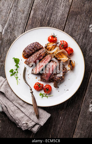 Fette di mezzo raro grigliate di carne di manzo Bistecca alla griglia con cipolle e pomodori ciliegia sulla piastra su sfondo di legno Foto Stock