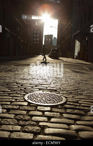 Silhouette di un uomo a piedi giù per una strada di ciottoli nel ponte di Brooklyn DUMBO quartiere. Focus su tombino in primo piano. Foto Stock