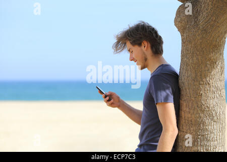 Vista laterale di un attraente uomo utilizzando un telefono intelligente sulla spiaggia con il mare e l'orizzonte in background Foto Stock