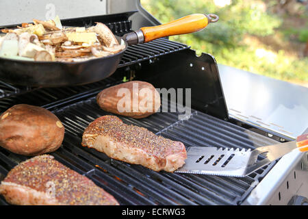 Una perfetta estate cena barbecue sotto il titolo sul grill, comprese bistecche, patate dolci e una padella di funghi e cipolle Foto Stock
