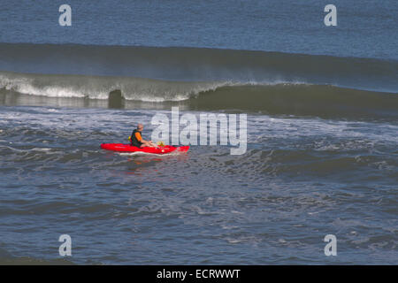 Un uomo in un rosso brillante e kayak giallo oar pale da un grande e la rottura delle onde oceaniche Foto Stock