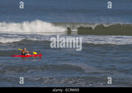 Un uomo in un rosso brillante e kayak giallo oar pale da un grande e la rottura delle onde oceaniche Foto Stock