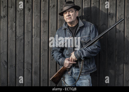 Uomo in cappello e camicia di jeans con un fucile da caccia di fronte ad una parete in legno guardando la telecamera. Foto Stock