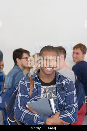 Studente maschio contenere libri e sorridente in telecamera con altri studenti in background Foto Stock