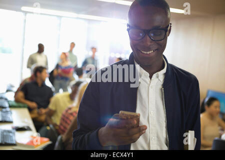 Sorridente studente maschio in occhiali guardando smart phone con altri studenti in background Foto Stock