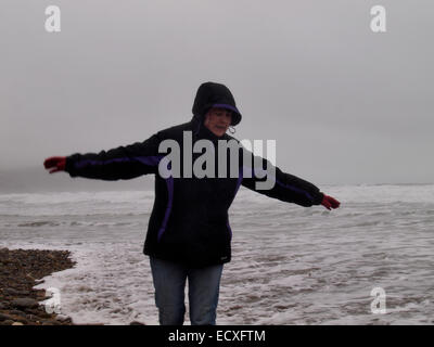 Donna in equilibrio su ciottoli di rimanere fuori dell'acqua sulla spiaggia in un grigio e nebbioso inverno di giorno, Widemouth Bay, Cornwall, Regno Unito Foto Stock