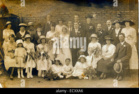 Circa 1920 Seppia pose foto di gruppo di famiglie estese in piedi intorno alla sposa e sposo dopo un matrimonio all'aperto con muro di mattoni dietro. Foto Stock