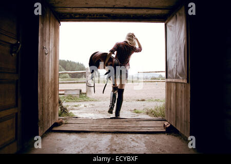 La donna caucasica porta sella di cavallo in fienile porta Foto Stock