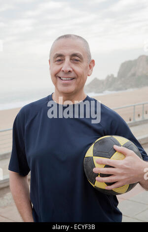 Senior ispanica uomo che porta palla calcio in spiaggia Foto Stock