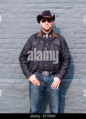 Un giovane uomo vestito da cowboy e indossando occhiali da sole pone in un modo maschilista da un muro di mattoni Foto Stock