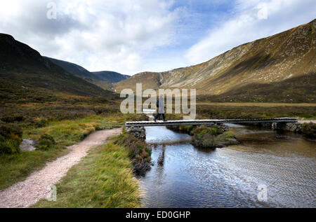 Una femmina di walker attraversa una passerella di legno sopra il Allt Un Dubh Loch con piste di bianco mese in background Foto Stock