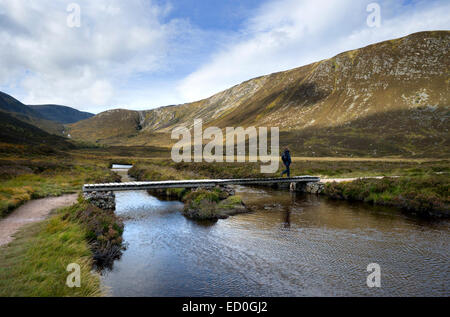 Una femmina di walker attraversa una passerella di legno sopra il Allt Un Dubh Loch con piste di bianco mese in background Foto Stock