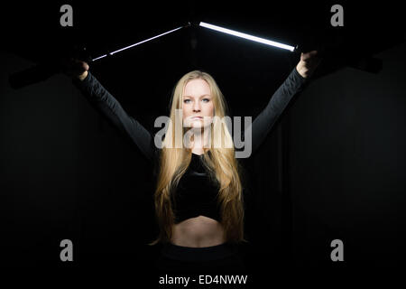 Un suggestivo forte eroico fiducioso giovane donna (18 anno vecchia ragazza) con lunghi capelli biondi di contenimento striscia due luci nel buio rivolta in avanti Foto Stock